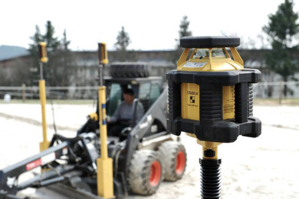 Detailansicht eines Nivelliergerätes, im Hintergrund Maschine zur Bearbeitung eines Reitplatzes