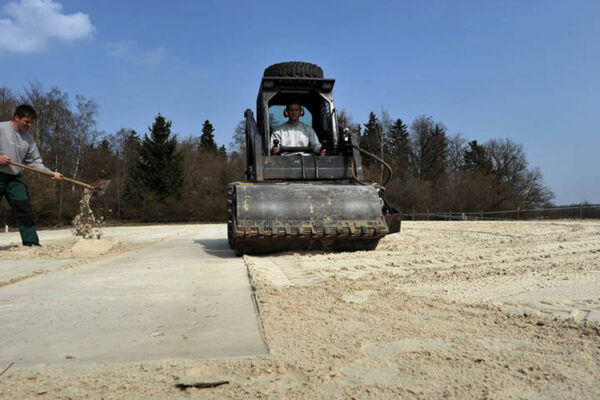 Arbeiter lockert Sand auf und Walze glättet die Oberfläche des Reitplatzes